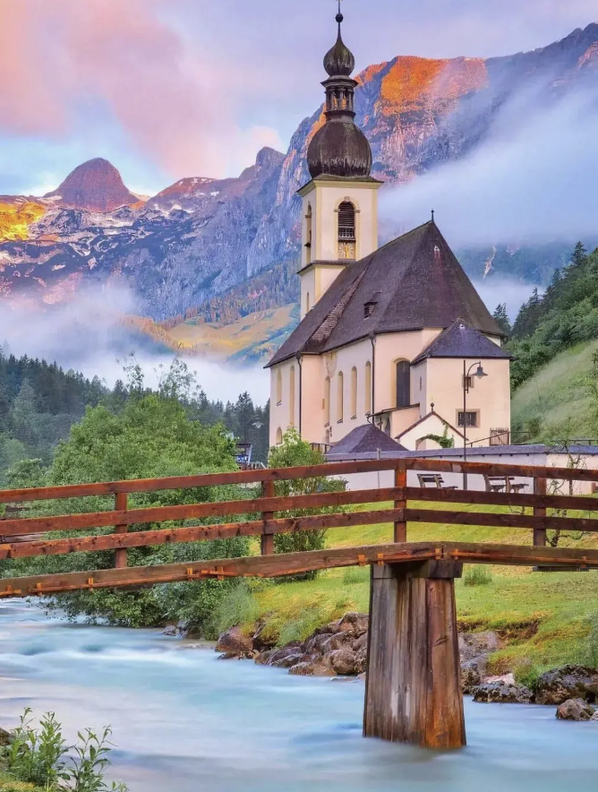 Une église au pied de montagnes baignées de brume, derrière un pont en bois et une rivière turquoise